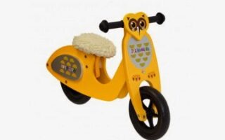 A yellow wooden balance bike for children with an owl face design on the front, black handle grips, a black front wheel, and a furry white padded seat. The side features a honeycomb pattern with small bees. The bike is against a plain white background.
