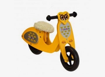 A yellow wooden balance bike for children with an owl face design on the front, black handle grips, a black front wheel, and a furry white padded seat. The side features a honeycomb pattern with small bees. The bike is against a plain white background.