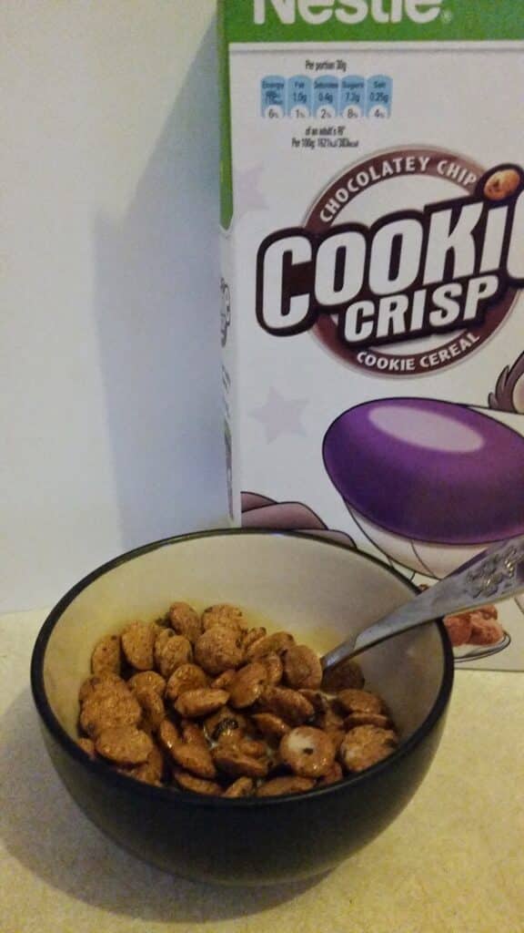A bowl of Cookie Crisp cereal with milk sits in front of a Cookie Crisp cereal box. A metal spoon is in the bowl. The box displays part of the brand name “Nestlé” and features purple and brown colours, with nutrition info visible at the top.