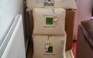 Three cream-coloured bedding items, each in protective bags with green and white labels and hang tags, are stacked against a white door. A radiator is on the left, and part of a brown sofa is visible on the right. The floor is light wood.