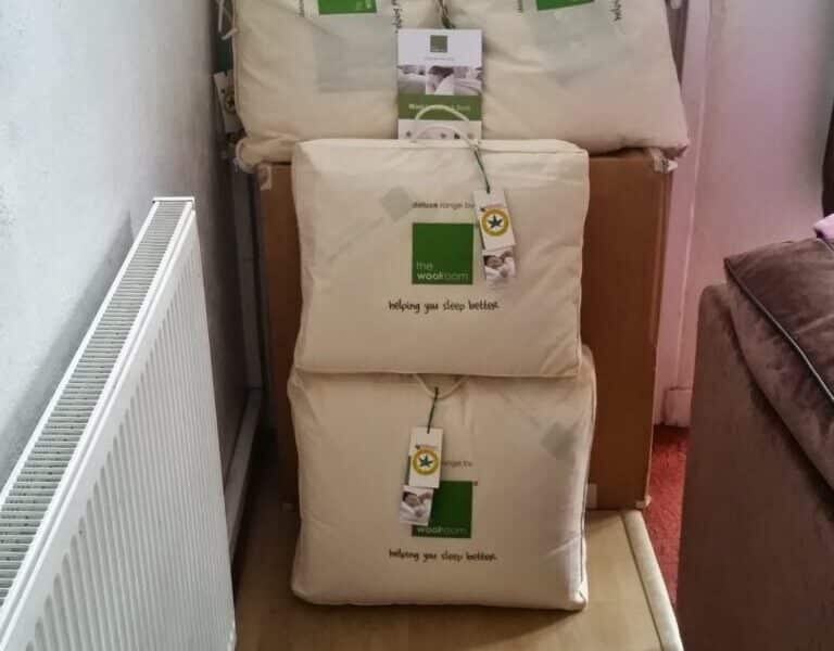 Three cream-coloured bedding items, each in protective bags with green and white labels and hang tags, are stacked against a white door. A radiator is on the left, and part of a brown sofa is visible on the right. The floor is light wood.