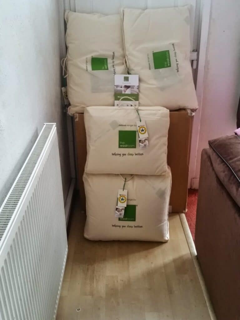Four cream-coloured bedding items, possibly duvets or pillows, are stacked against a white wall and radiator. Each item has a green and white label and a yellow tag, with the text "helping you sleep better" visible on the front packages.