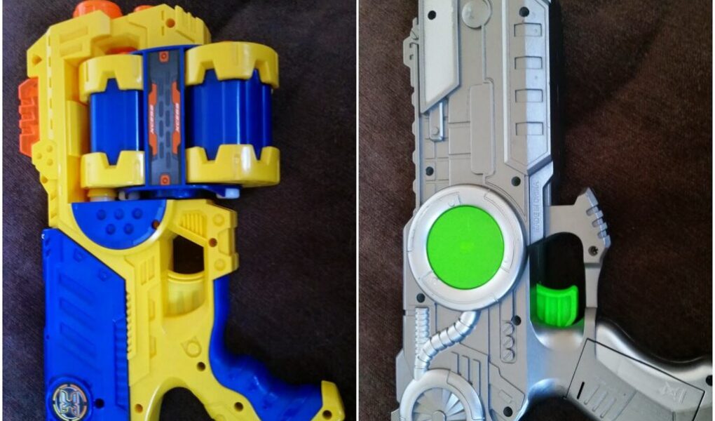 The image shows two colourful toy guns on a dark surface. The left toy is blue, yellow, and orange with a bulky, cylindrical chamber. The right toy is silver and grey with green accents, a sci-fi design, and a bright green trigger.
