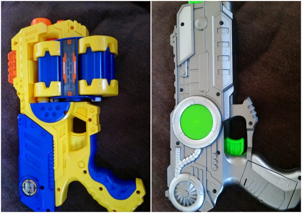 Side-by-side photo of two toy guns: left is blue and yellow with orange and a large blue cylinder; right is silver with green and black accents, futuristic design, and a round green detail above the trigger. Both are lying on a dark surface.