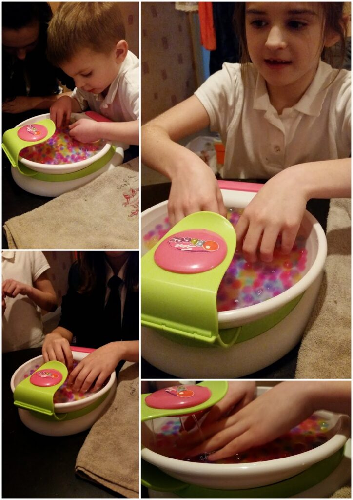 A collage shows two children playing with a bowl filled with colourful water beads, using their hands to mix and touch the beads. They look focused and happy, and a towel is placed nearby on a dark table for drying their hands.