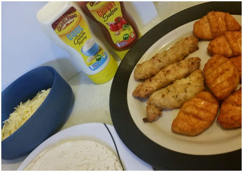 A plate of breaded chicken goujons and grilled chicken pieces is next to a blue bowl of grated cheese, a stack of flour tortillas, and bottles of Cool Chipotle Crema and Chunky Salsa sauces on a kitchen worktop.