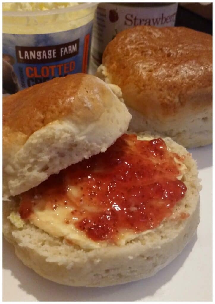 A close-up of two scones; one is sliced open and topped with clotted cream and strawberry jam, whilst the other is whole. In the background, a tub of clotted cream and a jar labelled "Strawberry" are partially visible.