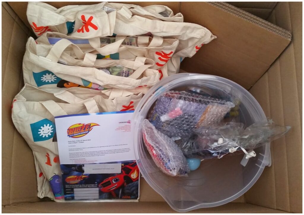 A cardboard box contains several canvas tote bags with colourful designs, a clear plastic bucket filled with small toys in plastic and bubble wrap, a toy car package, and a printed letter with a red and blue logo in the centre.