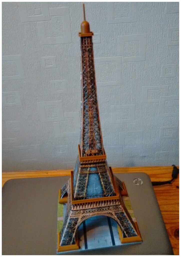 A small, detailed model of the Eiffel Tower stands on a closed HP laptop. The model shows structural details and is coloured to resemble the real tower. The background is a textured wall with geometric patterns, and part of a wooden desk is visible.