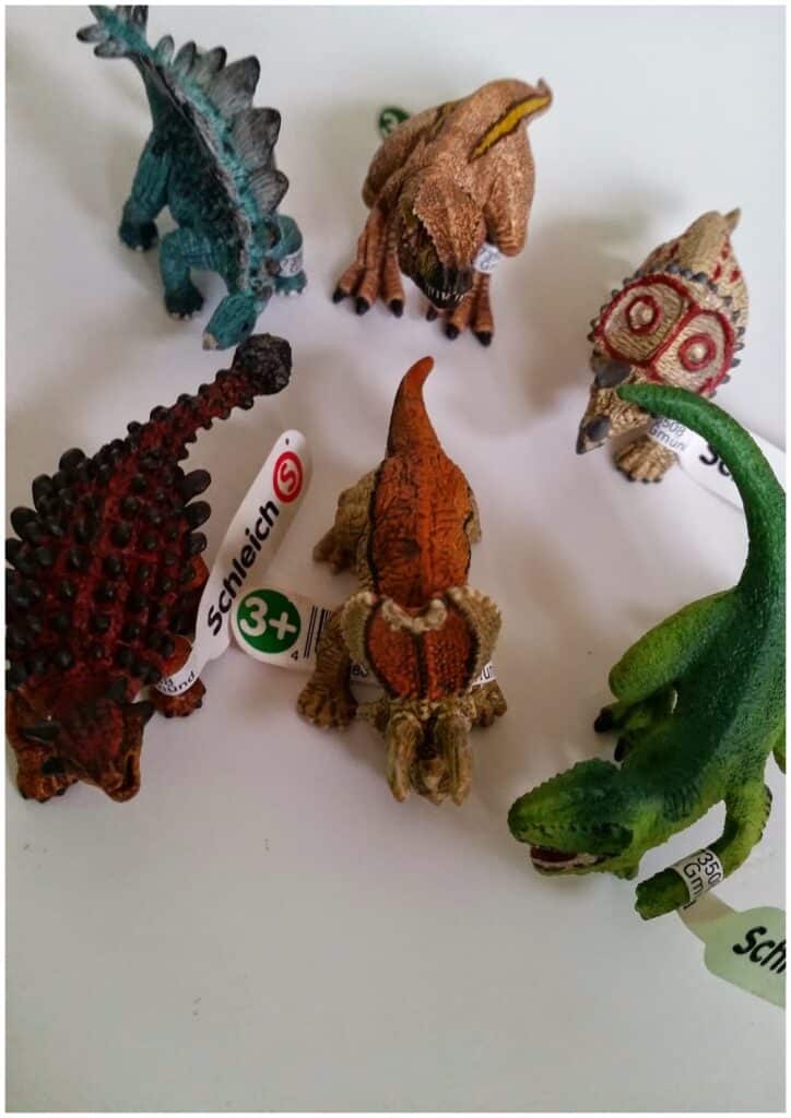 Six toy dinosaur figurines arranged in a loose circle on a white surface. Each is colourful and textured, with attached Schleich tags and a “3+” age recommendation visible. The dinosaurs vary in colour, markings, and posture.