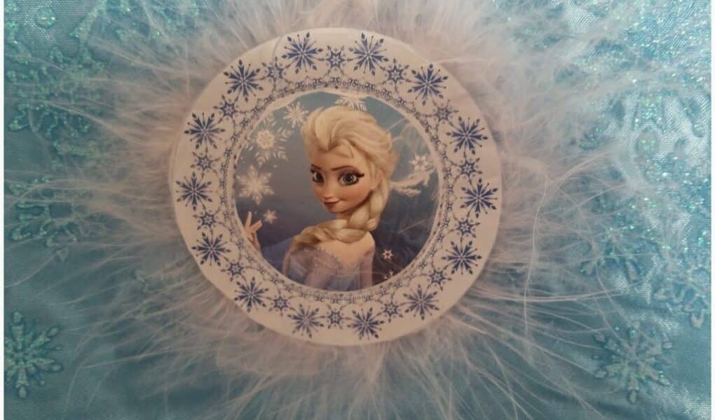 A circular ornament featuring Elsa from Disney’s Frozen, framed by blue snowflake patterns and surrounded by white feathery trim, is set against a sparkly light blue fabric with embroidered snowflakes.