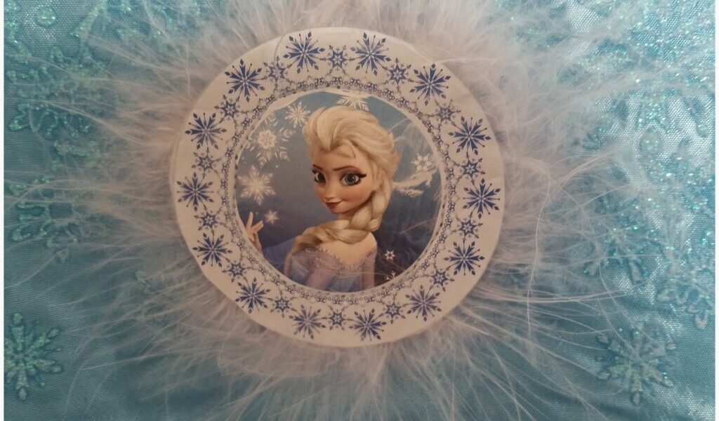A circular ornament featuring Elsa from Disney’s Frozen, framed by blue snowflake patterns and surrounded by white feathery trim, is set against a sparkly light blue fabric with embroidered snowflakes.