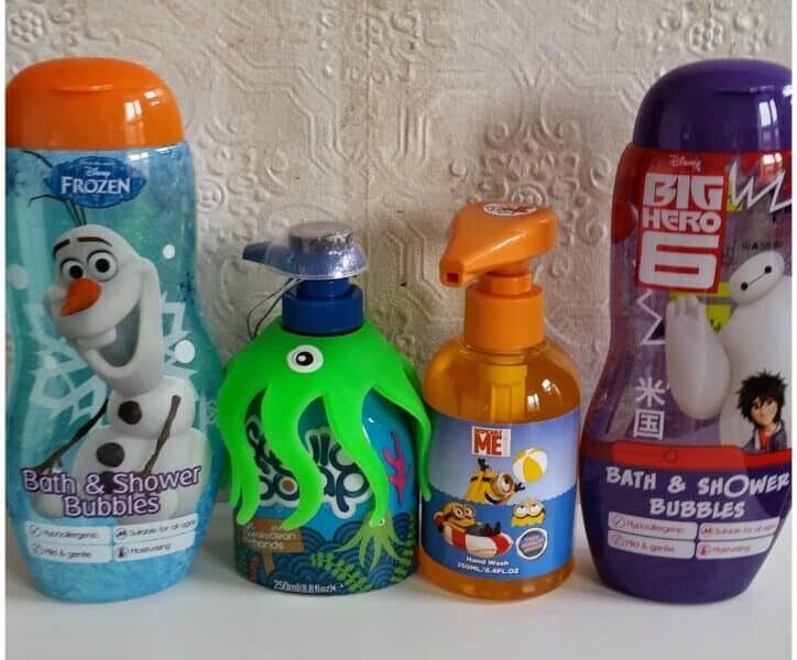 Four bottles of children’s bath products stand side by side. The first has Olaf from Frozen, the second is a green octopus-shaped bottle, the third is orange with Minions, and the fourth is purple with Baymax and Hiro from Big Hero 6. Textured white wall behind.