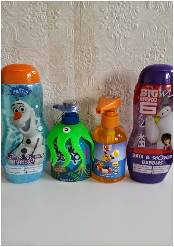 Four bottles of children's bath products stand in a row: a Frozen-themed bottle with Olaf, a green octopus-shaped foam soap dispenser, a Minions-themed pump bottle, and a Big Hero 6-themed bottle with Baymax and Hiro, set against a white textured wall.