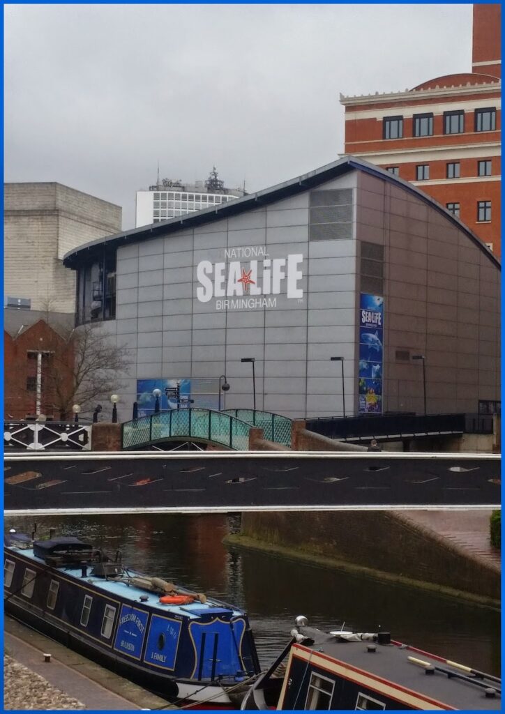 A view of the National SEA LIFE Centre in Birmingham, UK, with its modern glass and metal exterior and large logo. In the foreground, a canal with moored narrowboats runs alongside the building. Tall city buildings and cloudy skies are in the background.