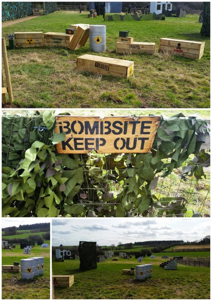 A paintball field with wooden crates labelled "Explosives," metal barrels with hazard symbols, and a "Bombsite Keep Out" sign on a green net fence. The field is grassy with obstacles and bunkers scattered throughout. Trees and hills are in the distance.