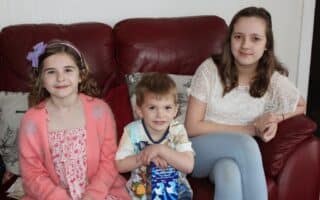 Three children sit on a red sofa. On the left, a young girl with wavy hair and a purple bow wears a pink dress. In the centre, a small boy in a colourful shirt holds a snack pack. On the right, an older girl in a white top and light blue trousers smiles.