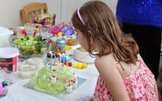 A young girl in a pink floral dress and headband leans forward to blow out candles on a green iced birthday cake decorated with small white rabbit figures. The table is filled with salads, snacks, and party food in a bright, indoor setting.