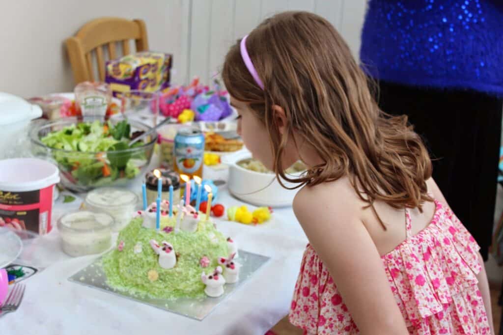 A young girl with light brown hair and a pink floral dress leans over a green birthday cake with lit candles, ready to blow them out. The cake is decorated with small bunny figures. The table is covered with party food and colourful decorations.