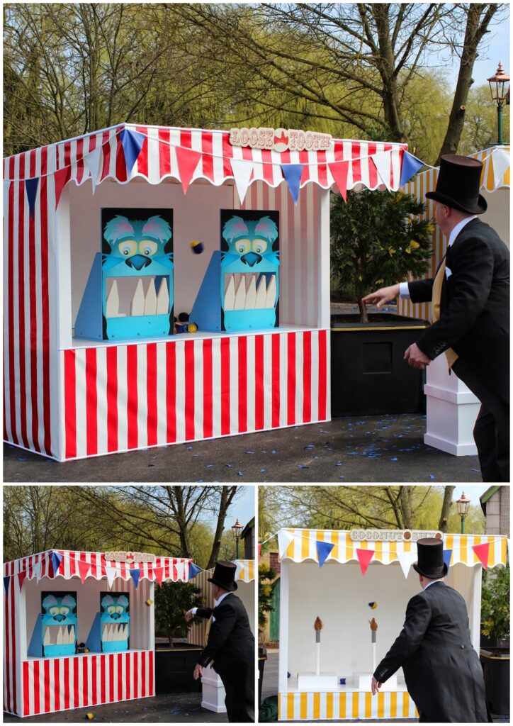 A carnival game stall with red-and-white stripes and blue, yellow, and red bunting. Two blue monster targets with big teeth face forwards. A man in a black suit and top hat throws balls at the game. Trees and lamp posts are in the background.
