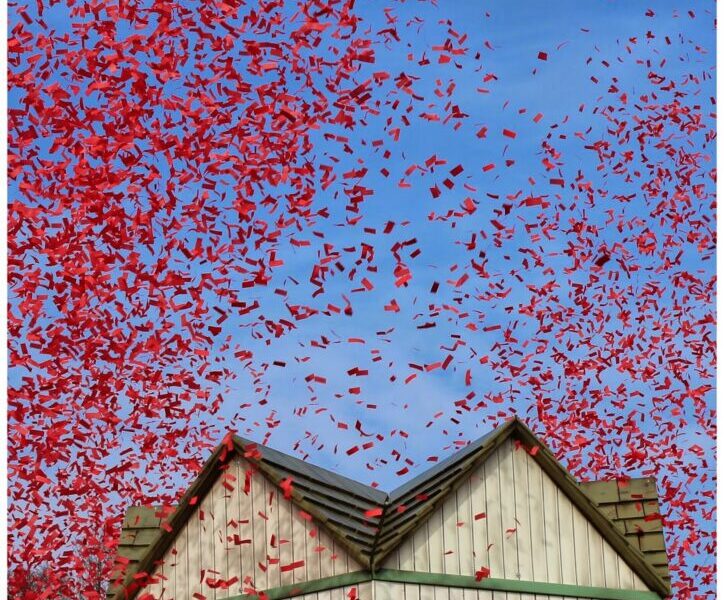 A flurry of red confetti fills the blue sky above a wooden structure with green trim and fake yellow-orange flames painted in the windows, suggesting a playful or festive event. The confetti swirls densely, creating a vibrant, celebratory scene.