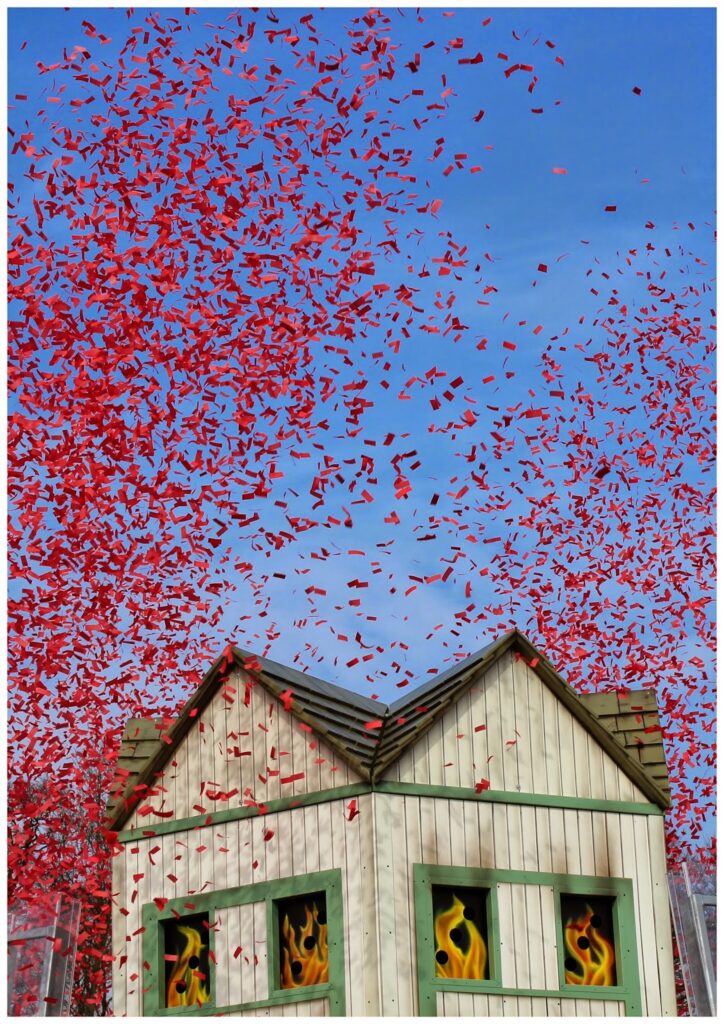 A small wooden house with green trim sits below a clear blue sky, whilst red confetti fills the air above. Yellow and orange flames appear in the windows, creating a vivid, festive, and dramatic scene.