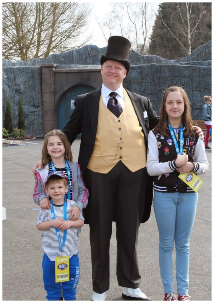 A man in a top hat and formal suit with a yellow waistcoat stands outdoors, smiling with three children. The children wear blue lanyards and are smiling. Trees and a stone wall are in the background, suggesting they are at a theme park or event.