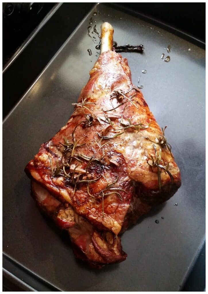 A roasted leg of lamb sits in a black baking tray. The meat is browned and covered with sprigs of fresh rosemary and pieces of roasted garlic, with some juices and oil visible around it. The lamb’s bone sticks out from one end.