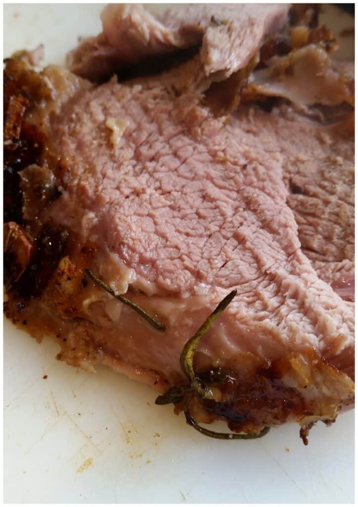 Close-up of a sliced piece of roast beef on a white chopping board, showing a browned, seasoned crust and tender, pinkish interior with visible texture and a sprig of rosemary.