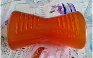 An orange, hourglass-shaped dog chew toy with grooves sits on a pastel, pastry-themed surface featuring images of cakes and cursive writing. The toy has the word "BIONIC" embossed on it.