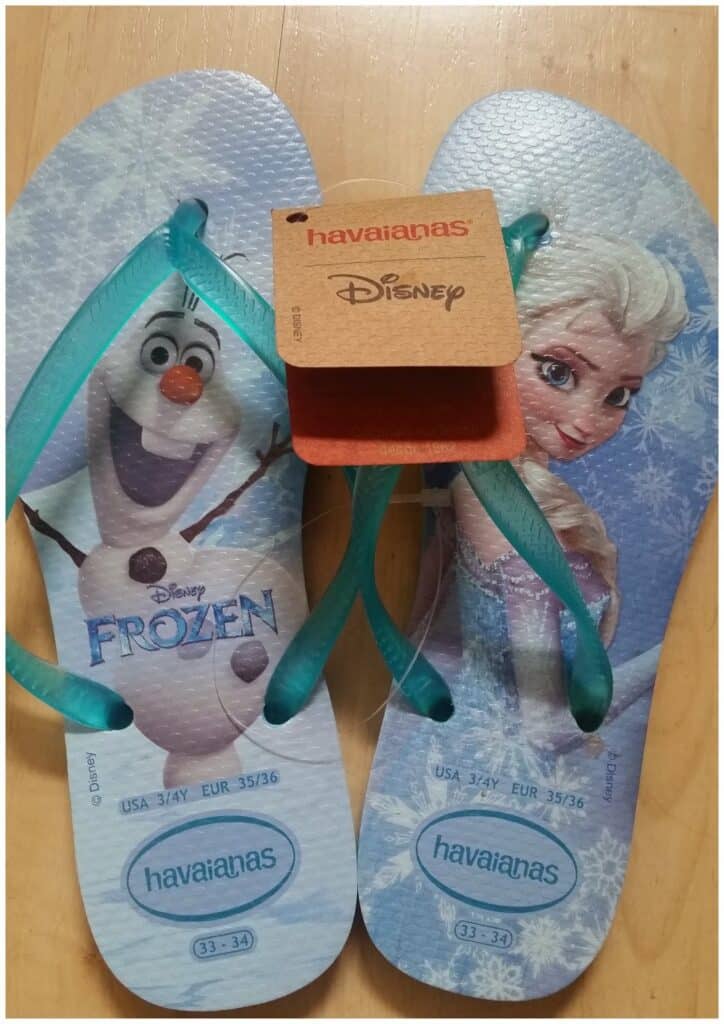 A pair of Havaianas flip-flops featuring Disney’s Frozen characters, Olaf the snowman on the left sandal and Elsa on the right, with blue straps. A brown Havaianas and Disney-branded tag is attached to the straps. The sandals are size 33-34.