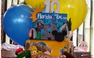 A colourful cake with theme park decorations, including the Universal Studios globe, E.T., Minions, Mickey Mouse ears, and Busch Gardens sign. The cake topper reads “10 FloridaTix.com.” Behind the cake are large yellow and blue balloons.
