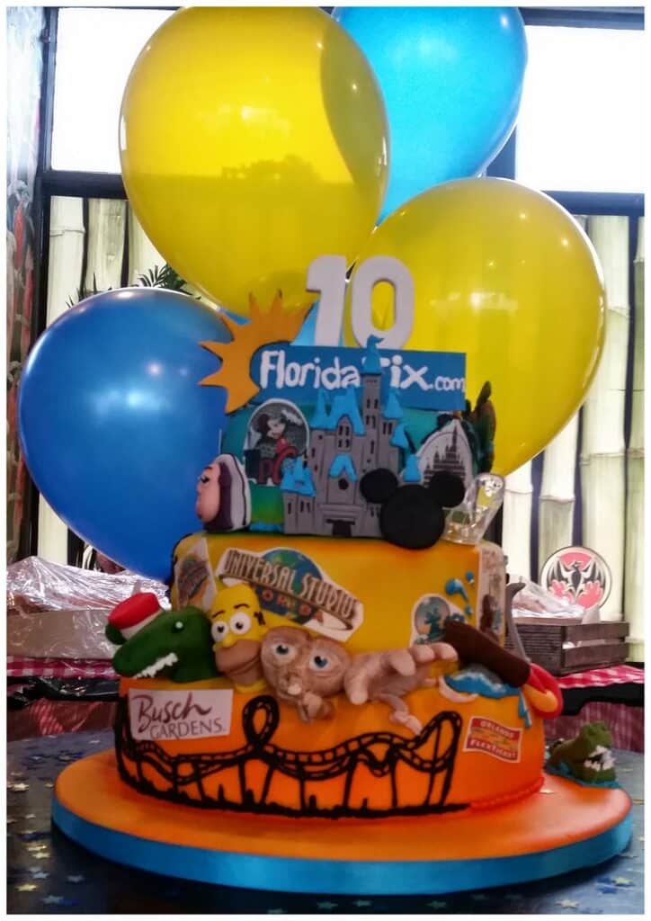 A two-tier cake decorated with Florida theme park icons, including Universal Studios, Busch Gardens, Mickey ears, E.T., and Simpsons characters. “10” and "florida-tix.com" top the cake. Blue and yellow balloons are in the background.
