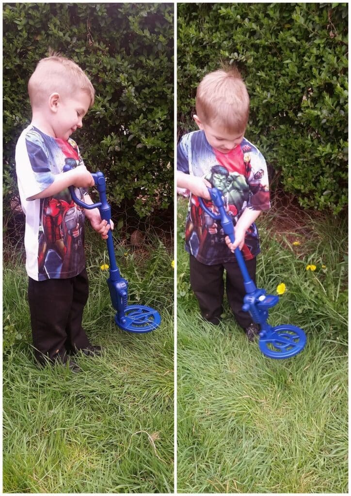 A young boy with short blond hair, wearing a superhero T-shirt and black trousers, stands on grass in front of green bushes. He holds a blue toy metal detector, exploring the grass in two different poses in a split image.