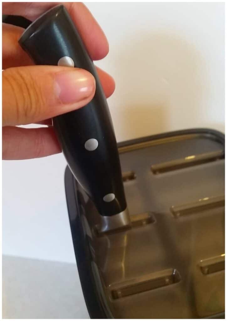 A hand holds the black, riveted handle of a kitchen knife above a black knife block with horizontal slots, ready to insert the knife. The background is white and minimalistic.