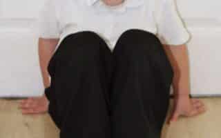 A young boy with short light brown hair sits on a light wood floor with his knees pulled up to his chest. He wears a white polo shirt, black trousers, and black shoes. His hands are placed behind him, and he looks up towards the camera with a neutral expression.