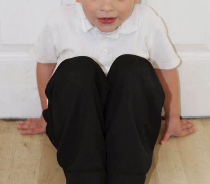 A young boy with short light brown hair sits on a light wood floor with his knees pulled up to his chest. He wears a white polo shirt, black trousers, and black shoes. His hands are placed behind him, and he looks up towards the camera with a neutral expression.