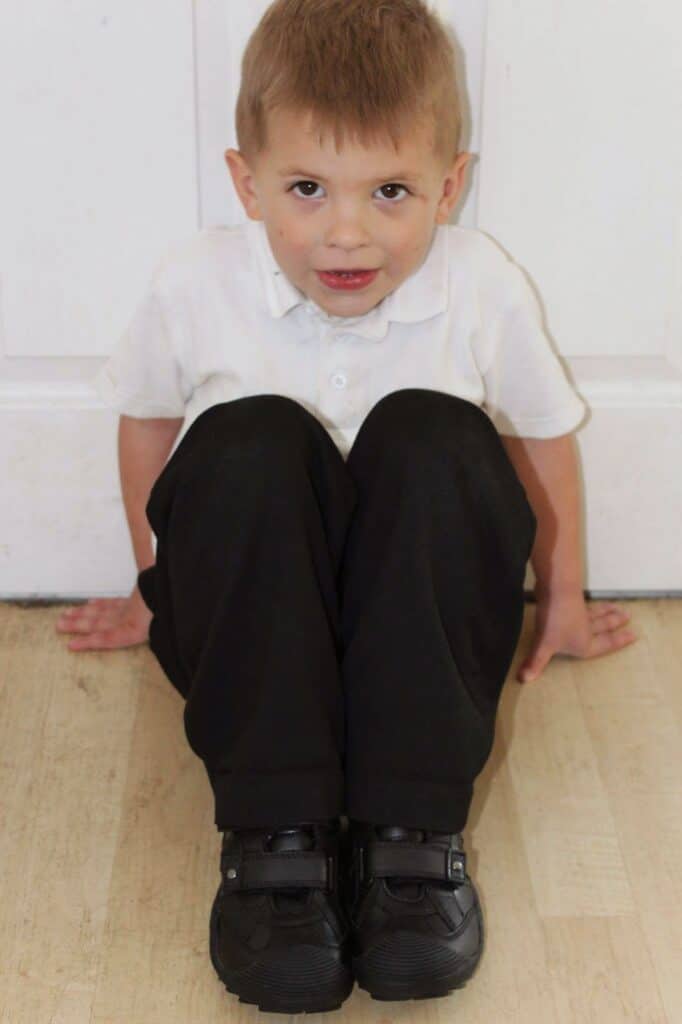 A young boy with fair hair, wearing a white polo shirt, black trousers, and black shoes, sits on a light wooden floor with his knees up and hands resting behind him. He looks slightly upwards, with a neutral expression, against a white wall background.