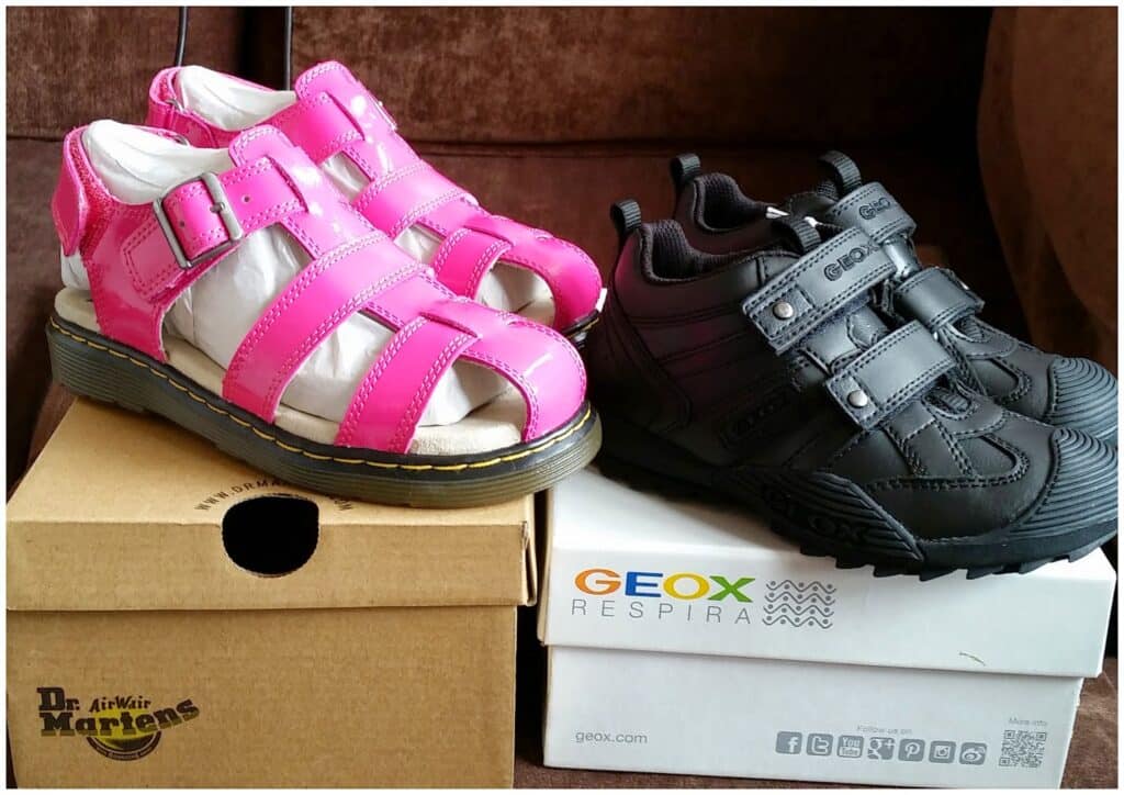Two pairs of children’s shoes sit on boxes: shiny pink Dr. Martens sandals with Velcro straps on a tan box, and black Geox Respira trainers with Velcro straps on a white box. Both boxes display their respective brand logos.