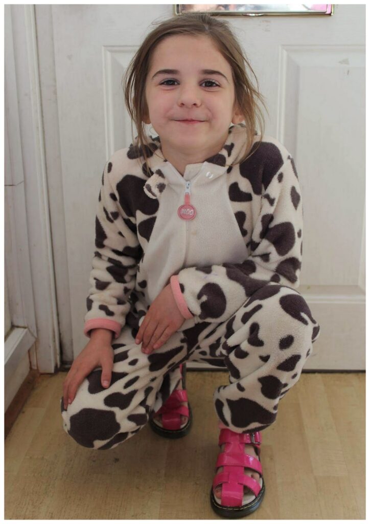 A young child with light skin and brown hair smiles while crouching indoors. They wear a cow-patterned onesie and bright pink sandals, posing in front of a white door on a light wooden floor.