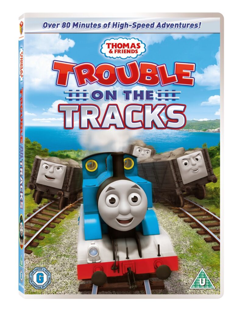 DVD cover for "Thomas & Friends: Trouble on the Tracks" shows Thomas the blue engine smiling on railway tracks, with three grey, surprised-looking train cars behind him. Green hills, blue sky, and a lake are in the background. Text reads “Trouble on the Tracks.”.