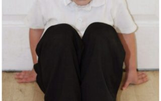 A young boy sits on a light wooden floor with his knees bent up to his chest. He wears a white polo shirt, black trousers, and black shoes. His hands are on the floor behind him for support, and he looks up at the camera. A white door is in the background.