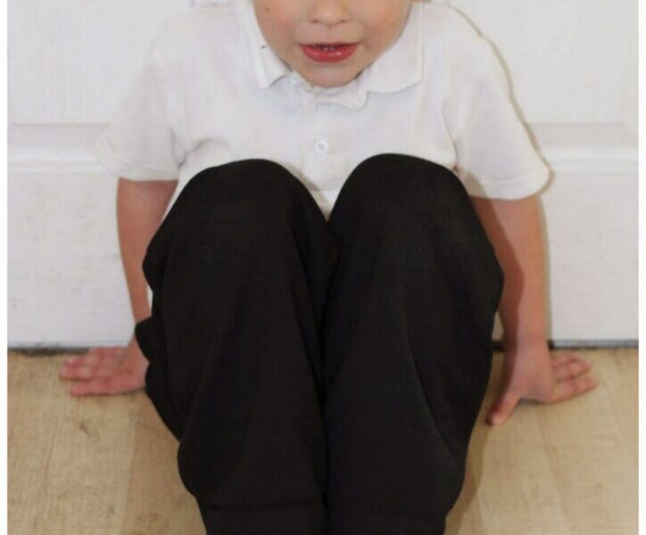 A young boy sits on a light wooden floor with his knees bent up to his chest. He wears a white polo shirt, black trousers, and black shoes. His hands are on the floor behind him for support, and he looks up at the camera. A white door is in the background.