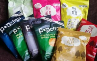 Nine colourful snack bags sit on a dark purple fabric background. Six are Ten Acre brand crisps in yellow, pink, green, and red, and three are Popchips in blue, green, and navy. Each bag displays its flavour and logo prominently.