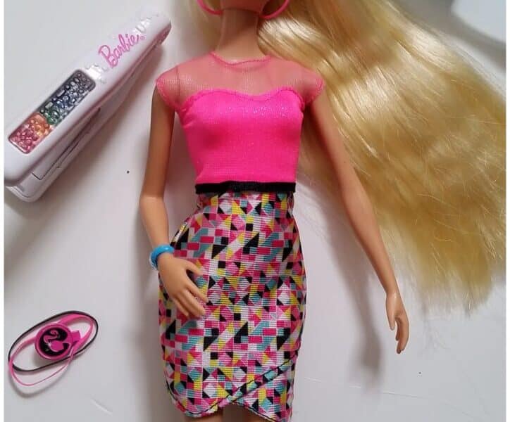 A Barbie doll with long blonde hair, wearing a bright pink top and a colourful geometric-patterned skirt, lies on a white surface. Around her are a blue brush, pink bracelet, Barbie-branded case, and assorted small hair accessories.