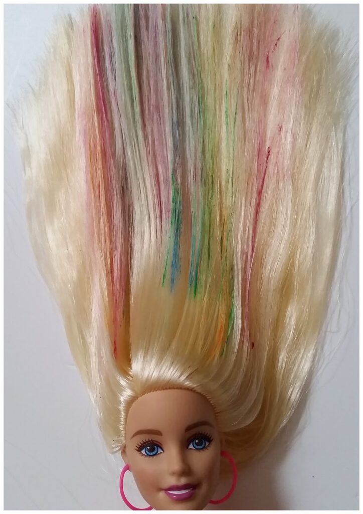 A blonde doll's head with blue eyes and pink hoop earrings is shown upside down. Her long hair is spread out above her, featuring streaks of various colours, including green, red, and blue, against the blonde strands. The background is plain white.