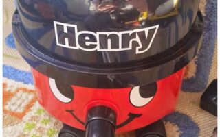 A red Henry vacuum cleaner with a smiling face and large eyes is on a patterned carpet. Its black hose is attached at the front, and the name “Henry” is written in bold white letters on the black top. Reflections are visible on the shiny surface.