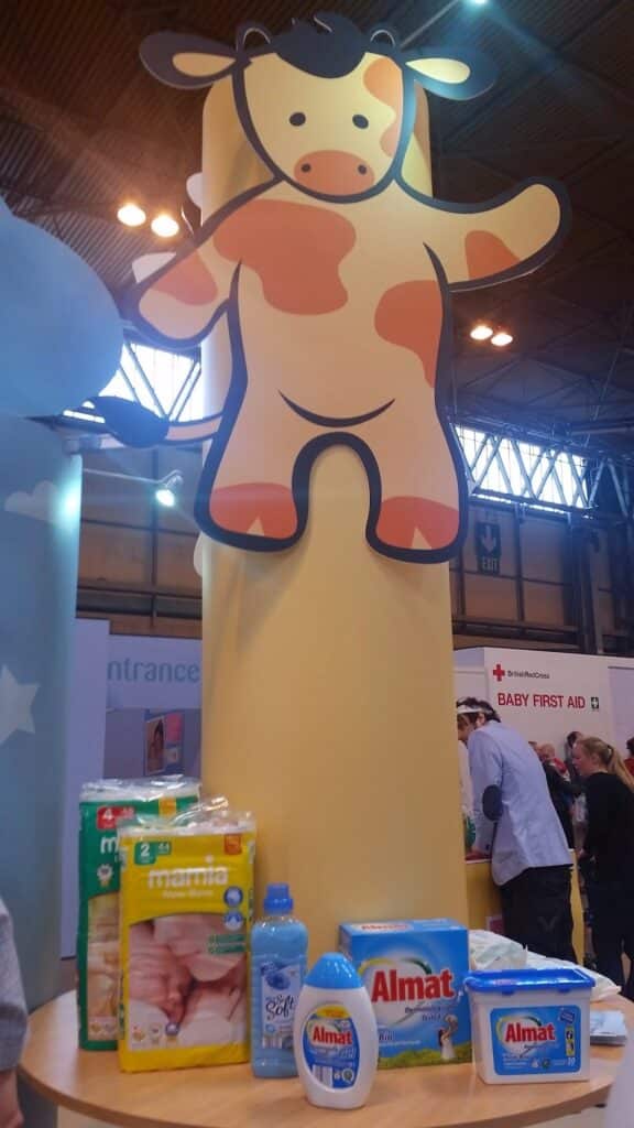 A large yellow pillar features a cute cartoon cow with orange spots and a smiling face. In front of the pillar, a table displays baby products including nappies, baby wipes, washing liquid, and fabric softener. People and stands are visible in the background.