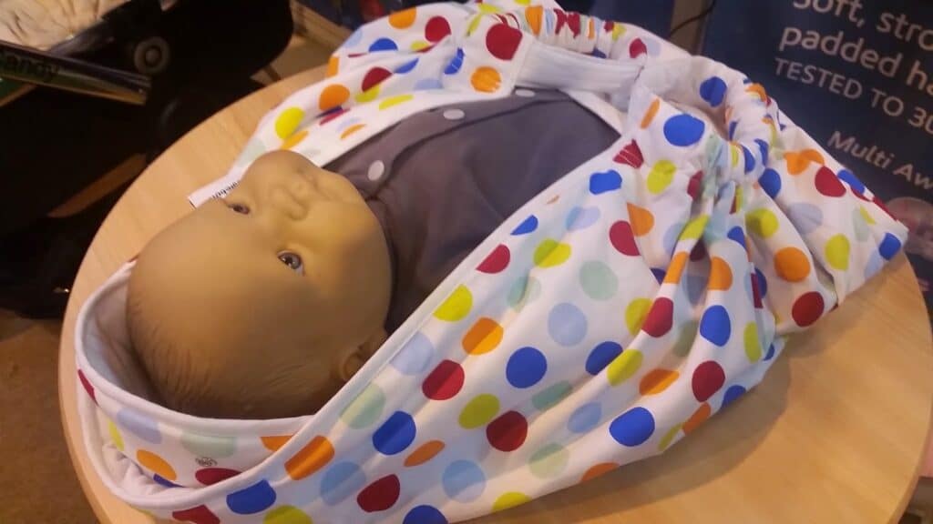 A lifelike baby doll wrapped in a white blanket with colourful polka dots lies on a round wooden table. The doll wears a grey outfit and looks upward. The scene appears to be in an indoor setting, possibly a display or demonstration.