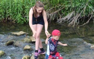 A girl in a black outfit helps a younger boy in a red cap and shorts cross a shallow stream by holding his hand as he steps on stones, surrounded by green grass and plants. The boy looks focused, and the girl is supporting him from behind.
